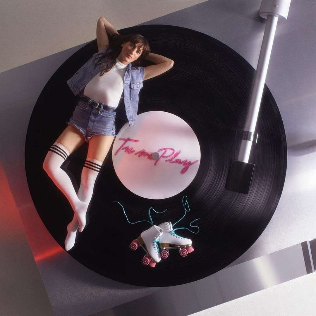 Woman lying on a record player next to roller skates, wearing a denim jacket and shorts, and knee-high socks with stripes.