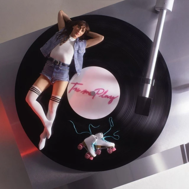 Woman lying on a record player next to roller skates, wearing a denim jacket and shorts, and knee-high socks with stripes.