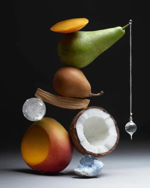 Stacked fruit arrangement featuring a mango, pear, and coconut with a crystal ball and metal pendulum against a dark grey background.