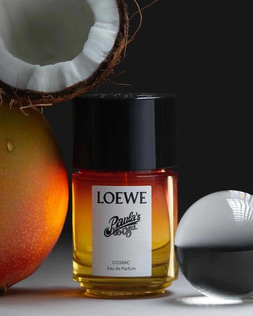Loewe Paula's Ibiza Cosmic perfume bottle stands near coconut and mango against a dark backdrop in a beauty product shot.