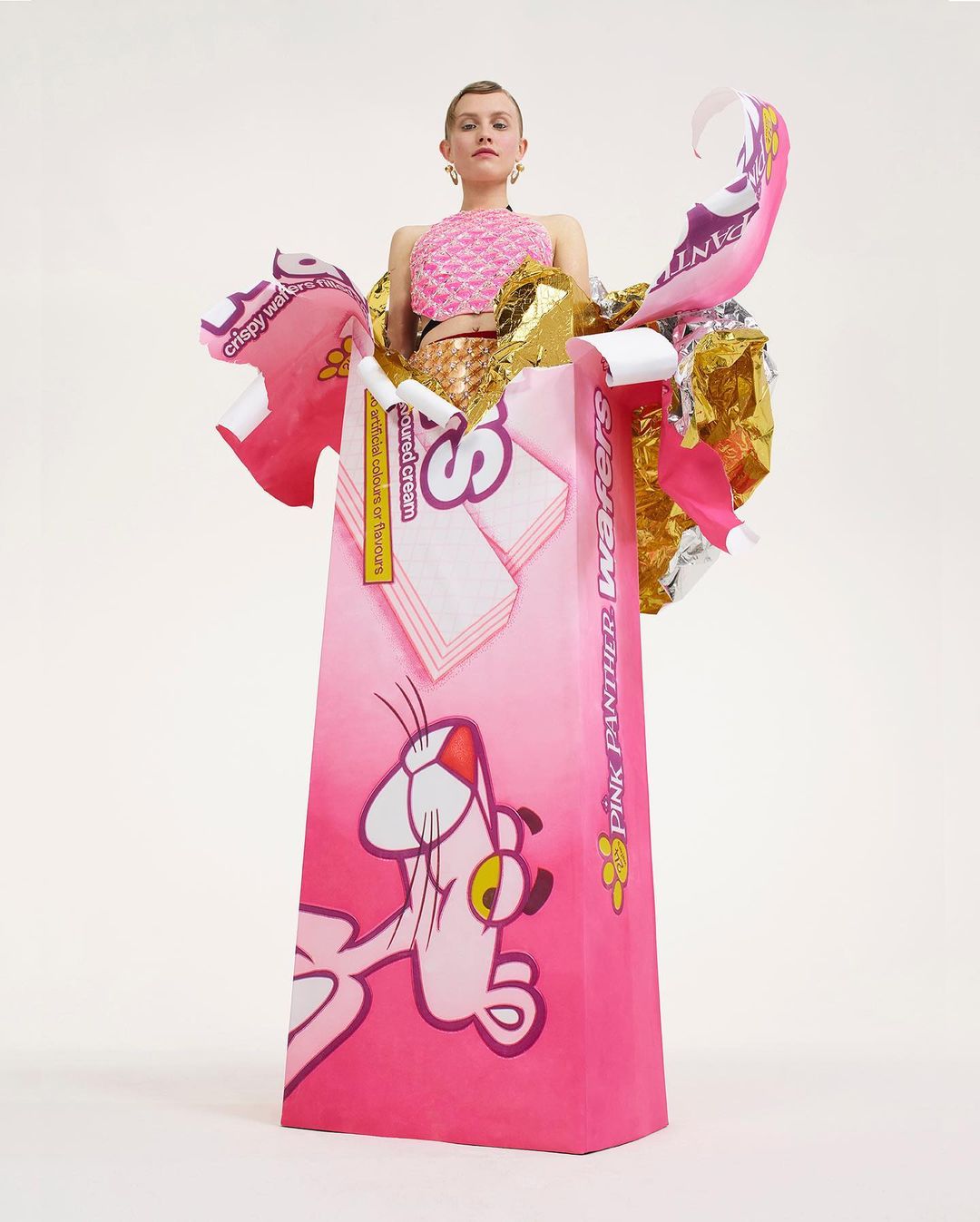 Adult woman stands in a pink Pink Panther Waters candy wrapper dress with exposed midriff in a studio setting on a white backdrop.