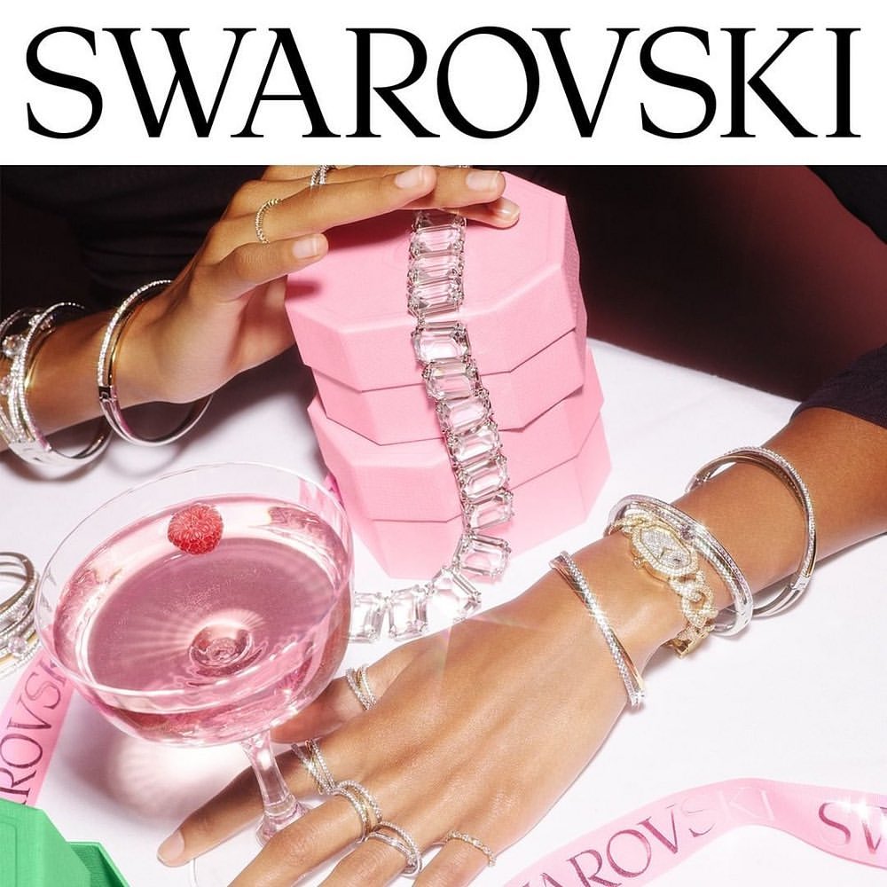 Close-up shot of jewelry by Swarovski, including rings, bracelets, and a necklace, displayed with a pink gift box and a cocktail.