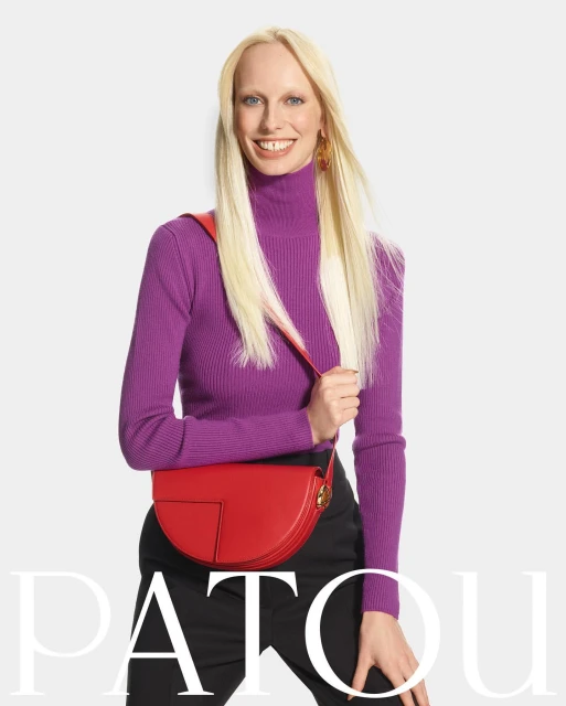 Smiling fair-skinned woman wearing a purple turtleneck and black pants, modeling a red handbag in a fashion studio.