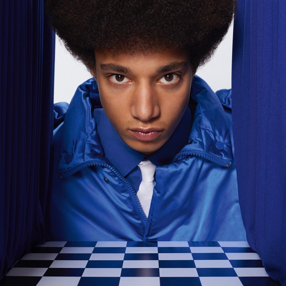 Stylish adult male with an afro, wearing a blue jacket and white tie, stands in front of a blue curtain and checkered floor.