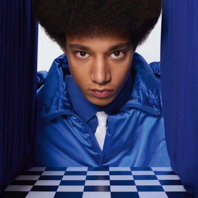 Stylish adult male with an afro, wearing a blue jacket and white tie, stands in front of a blue curtain and checkered floor.