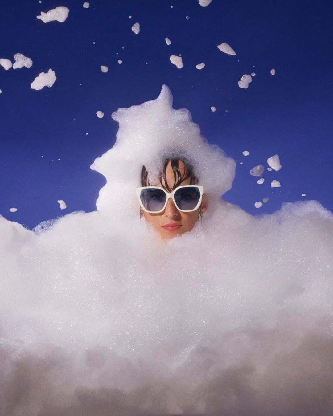 Eye-level shot of an adult female wearing sunglasses, partially submerged in white foam against a royal blue background in a studio.