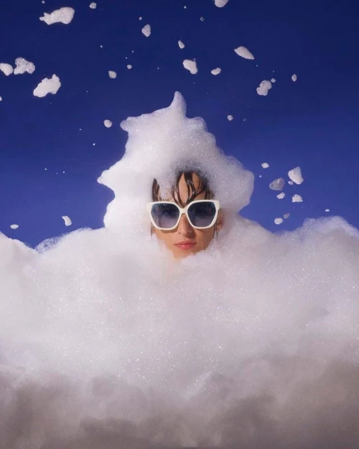 Eye-level shot of an adult female wearing sunglasses, partially submerged in white foam against a royal blue background in a studio.