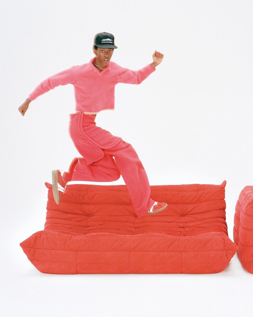 An adult male is jumping over a red Togo sofa in a photo studio. He wears a black cap, red sweater and red pants against a white background.