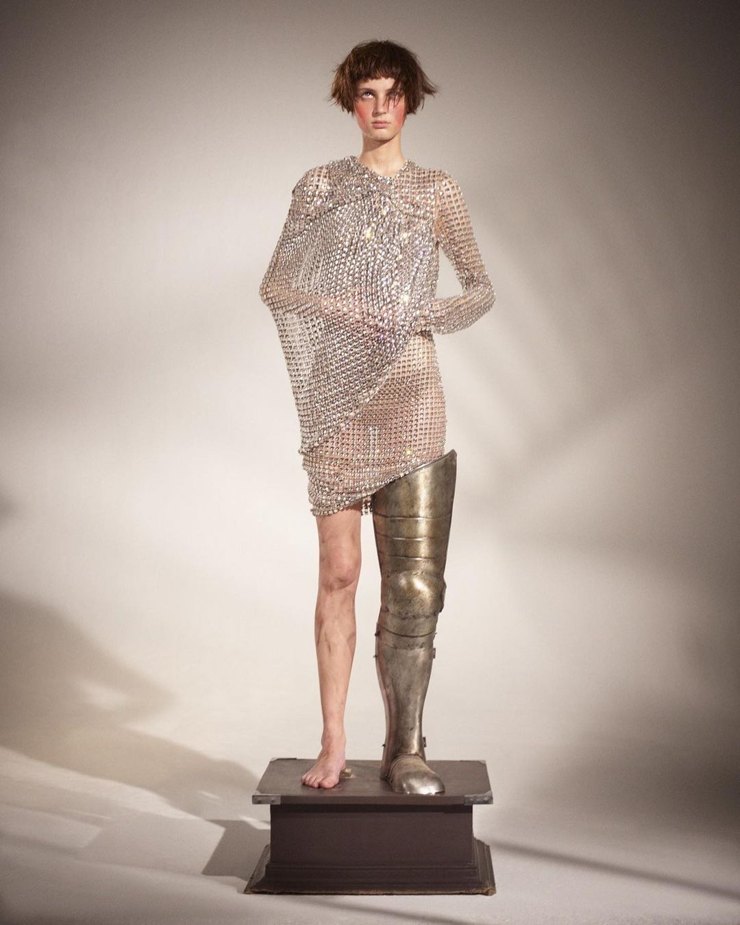 Fashionable person stands on a wooden platform wearing a metal chainmail dress and knight's leg armour, studio lighting, neutral backdrop.