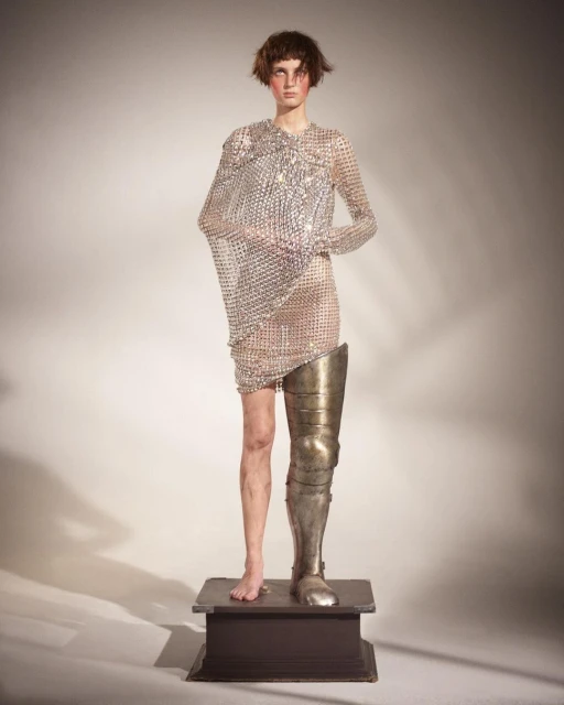 Fashionable person stands on a wooden platform wearing a metal chainmail dress and knight's leg armour, studio lighting, neutral backdrop.