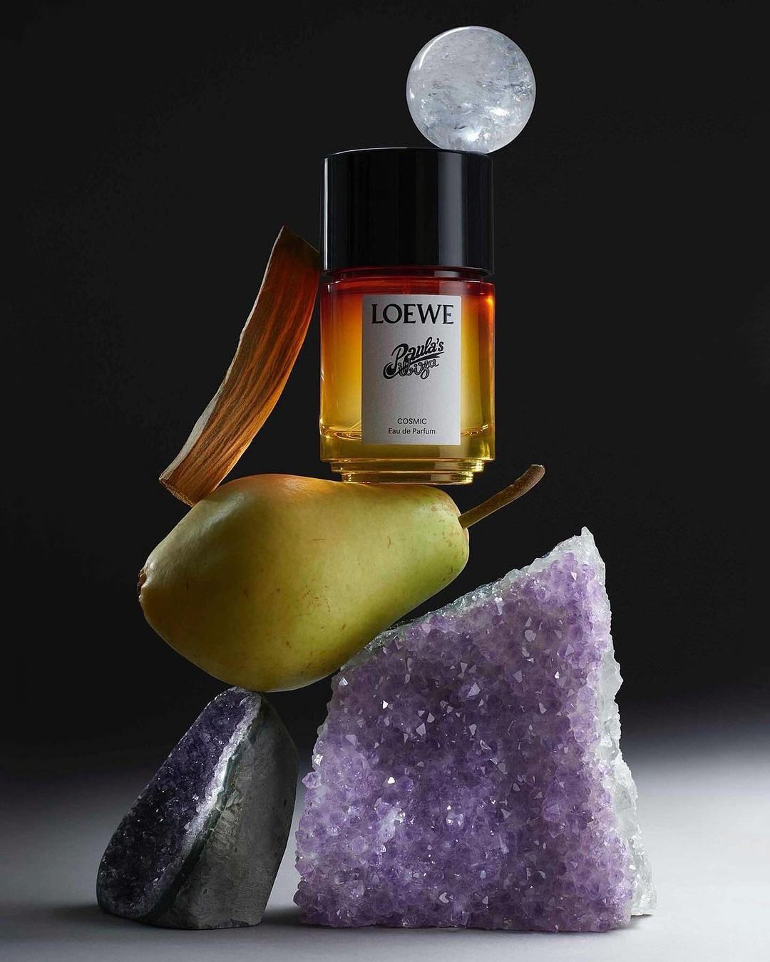 Still life of Loewe Esencia EdP perfume bottle balanced with a pear, amethyst, clear crystal, and dried leaf against dark background.