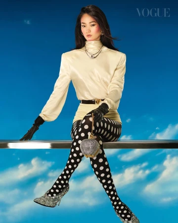 A stylish Asian woman poses in a satin blouse, polka-dot leggings, and glittering heels against a blue backdrop for Vogue magazine.