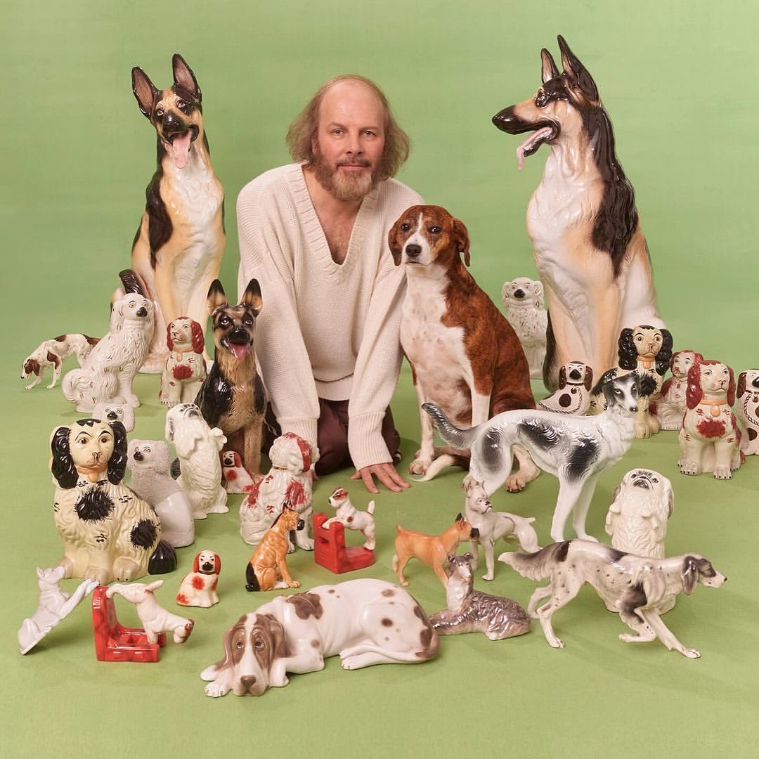 Man with a beard surrounded by many ceramic dog figurines of various breeds in a studio setting with a light green background.
