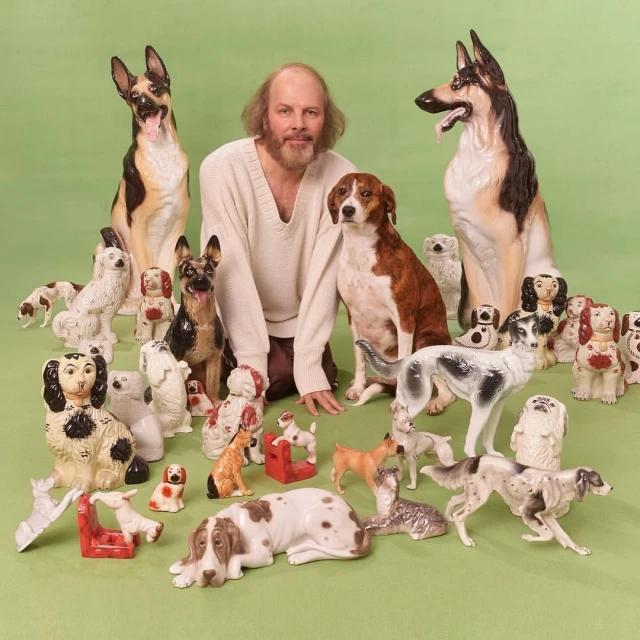 Man with a beard surrounded by many ceramic dog figurines of various breeds in a studio setting with a light green background.