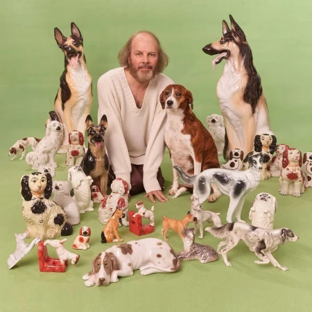 Man with a beard surrounded by many ceramic dog figurines of various breeds in a studio setting with a light green background.