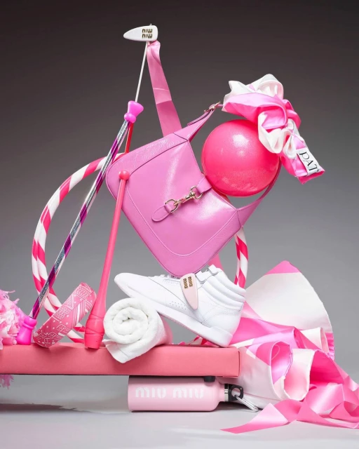 Pink-themed still life featuring a handbag, sneakers, gymnastic ball, water bottle, and ribbons, creatively arranged on a pink platform.