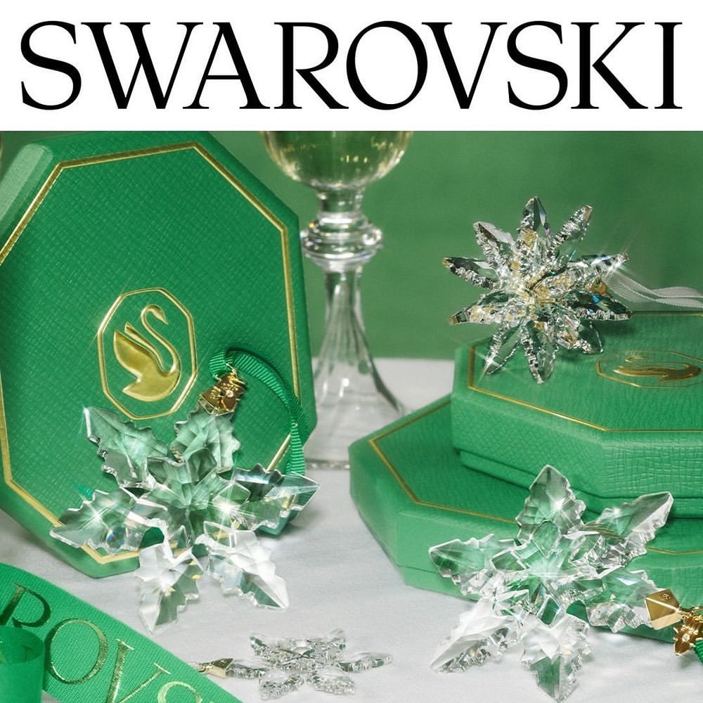 Crystal snowflake ornaments and green gift boxes arranged against a green backdrop, highlighted by a stemmed glass and Swarovski branding.