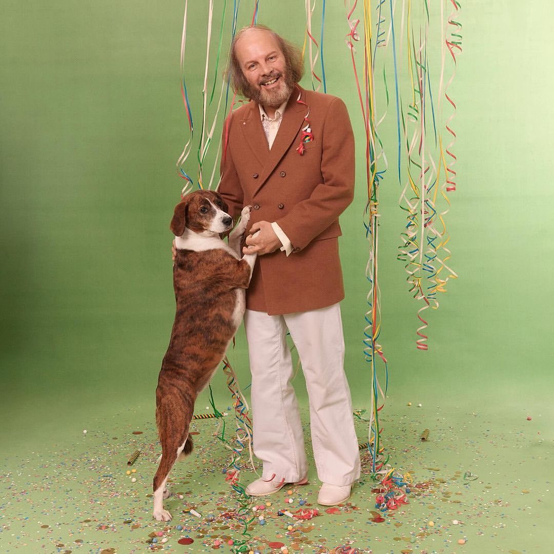 A man in a brown suit poses with a brown and white dog in a green photo studio with confetti and streamers, looking happy.