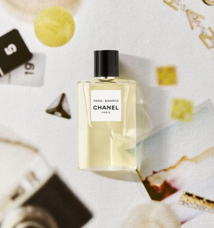 A rectangular Chanel Paris Biarritz perfume bottle resting on a textured, flat lay surface featuring assorted cards and papers.