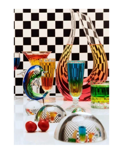 Still life of colorful liquid filled glassware including stemware, tumblers, and a dolphin figurine against a checkered backdrop.