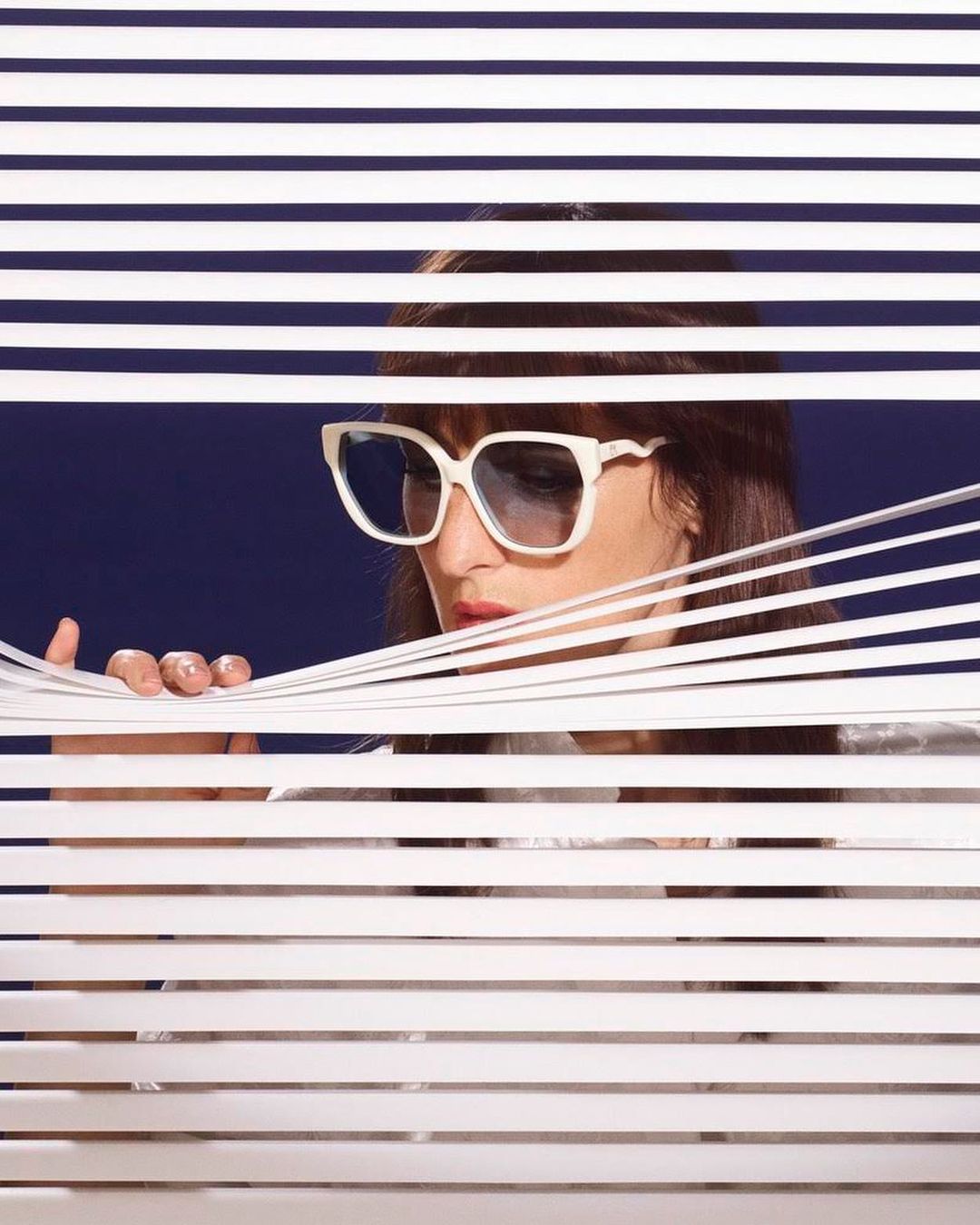 Woman with dark hair and large white sunglasses peeks through horizontal blinds against a dark blue background in a studio portrait.