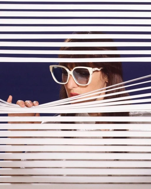 Woman with dark hair and large white sunglasses peeks through horizontal blinds against a dark blue background in a studio portrait.