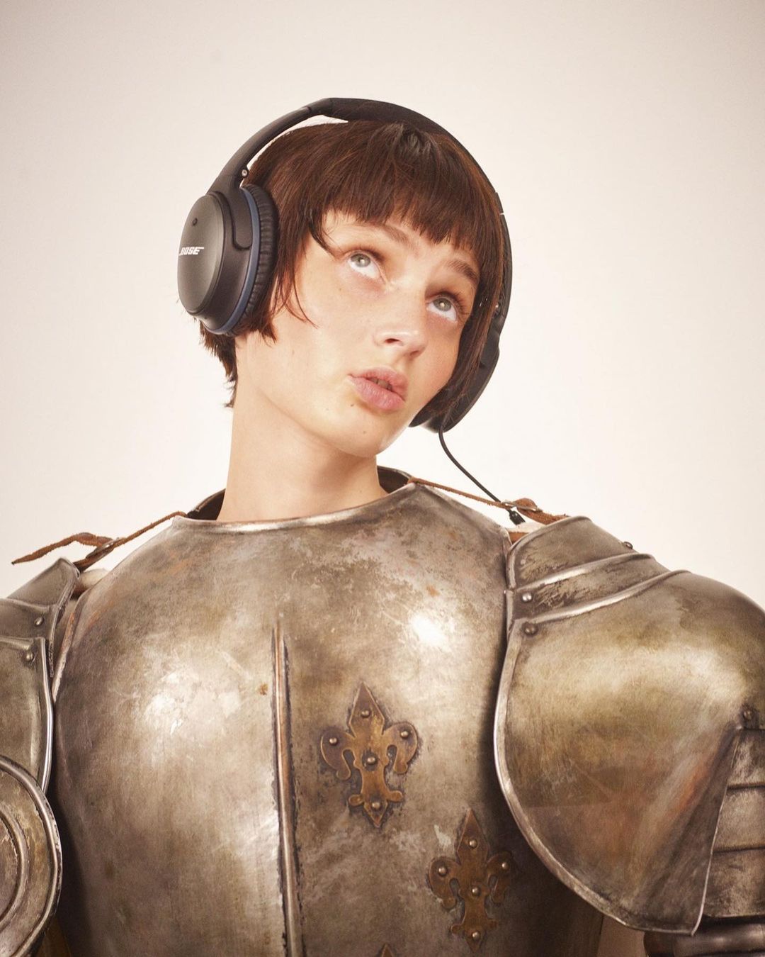 Portrait of a young adult in armour and headphones looking up, against a neutral background. Conceptual portrait.