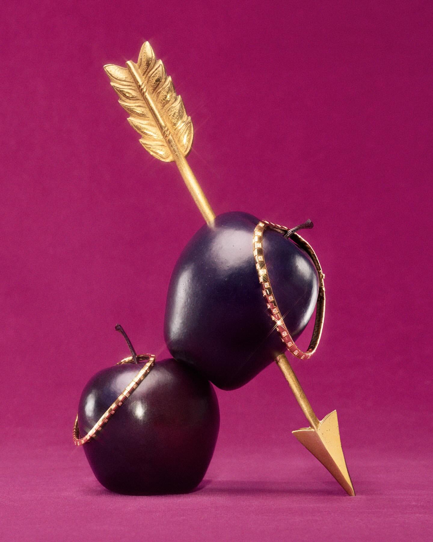 Still life of two dark apples stacked, pierced by a gold arrow, adorned with a gold chain, against a magenta background.