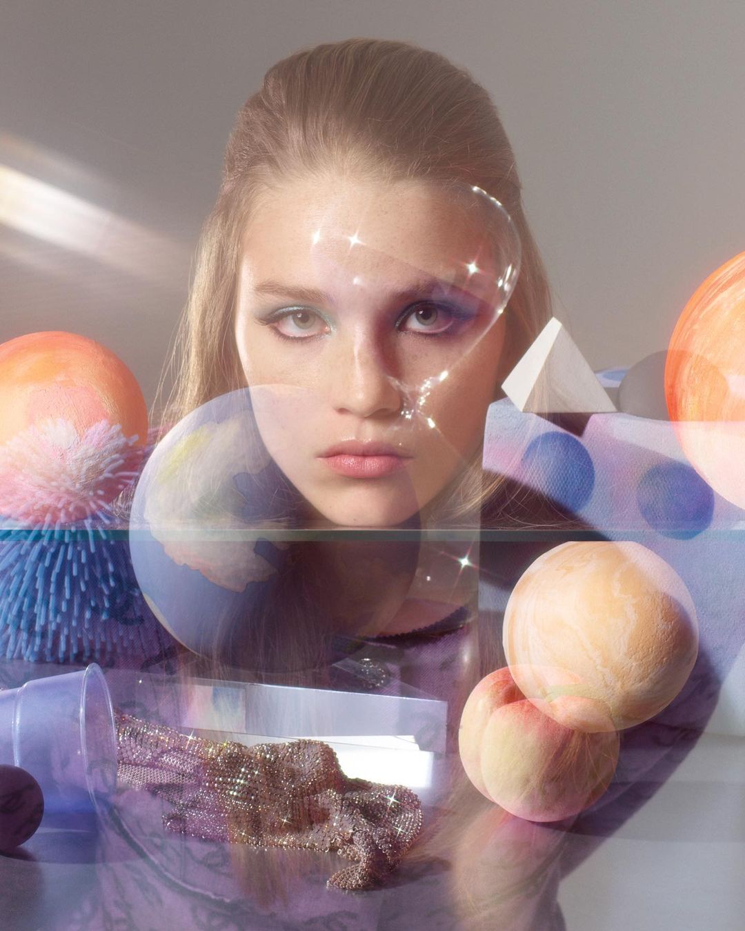 Portrait of a young woman with makeup, illuminated by diffused lighting, surrounded by spheres, a pyramid, and a scattering of glitter.