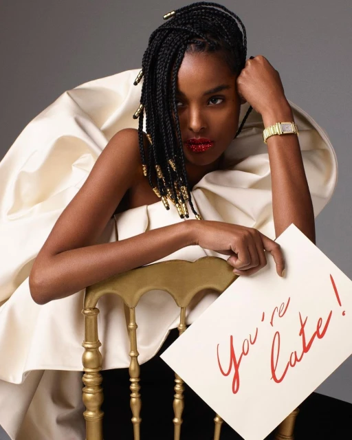 Elegant portrait of a Black model in a white ruffle dress sitting on a gold chair holding a 'You're late!' card in a grey studio.