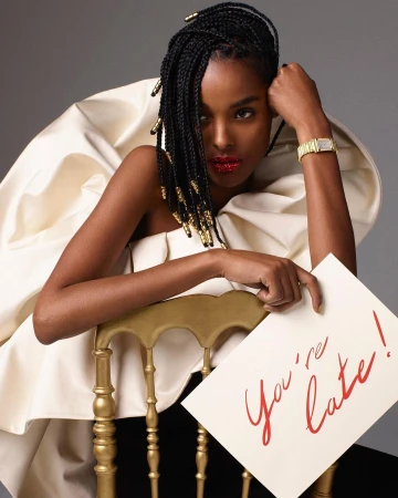 Elegant portrait of a Black model in a white ruffle dress sitting on a gold chair holding a 'You're late!' card in a grey studio.