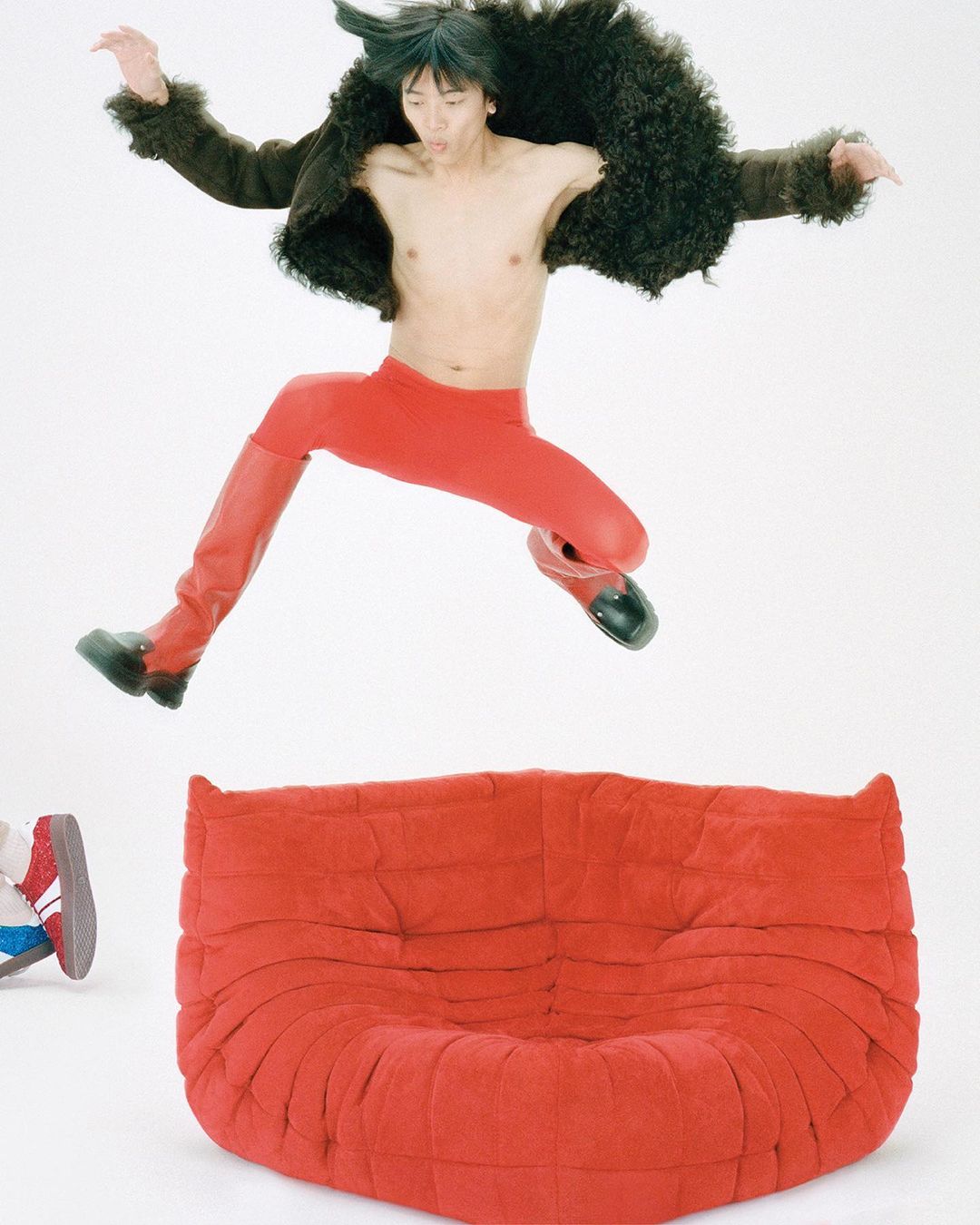 An Asian man in a fur jacket and red leggings jumps high in the air near a red sofa, capturing a lively and dynamic fashion scene.