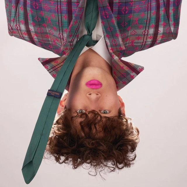 Young adult male with pink lipstick, wearing a plaid jacket and green tie, photographed in an upside-down orientation against a gray background.
