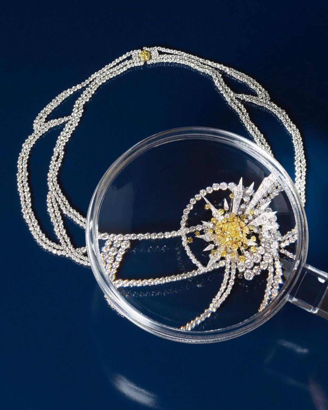 Close-up shot of an elaborate diamond necklace with a magnifying glass, set against a dark blue background, highlighting its details.