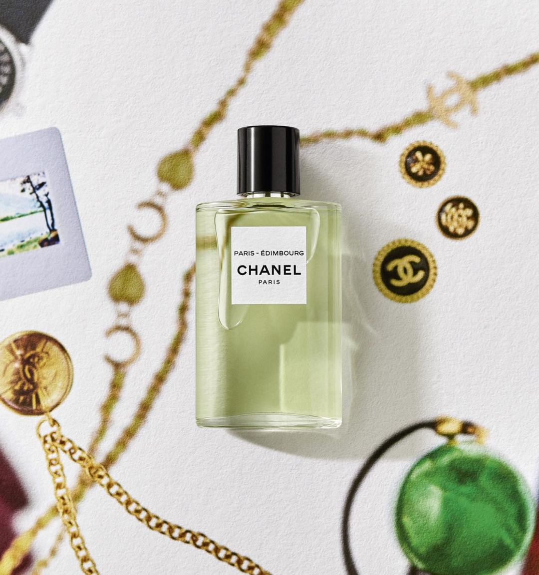 Still life of Chanel Paris-Edimbourg perfume bottle with gold chains, vintage buttons, and a photo on a textured white surface.