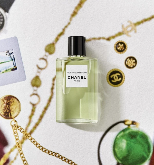 Still life of Chanel Paris-Edimbourg perfume bottle with gold chains, vintage buttons, and a photo on a textured white surface.
