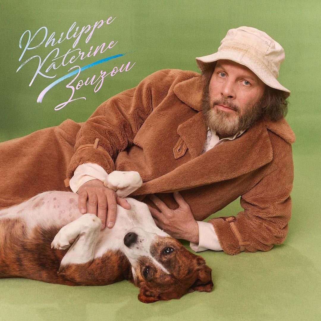 Adult male and dog lying on green surface. Man wears a brown coat and light-colored hat. The dog is brown and white with its belly exposed.