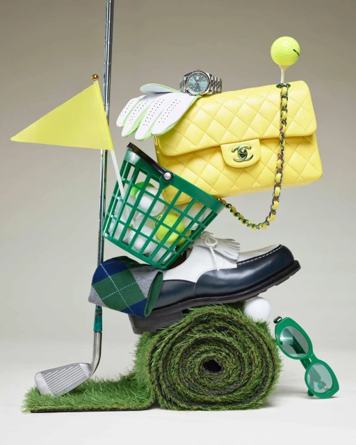 Still life of golf equipment and luxury goods stacked on artificial turf, featuring a golf club, golf balls, handbag, and watch.