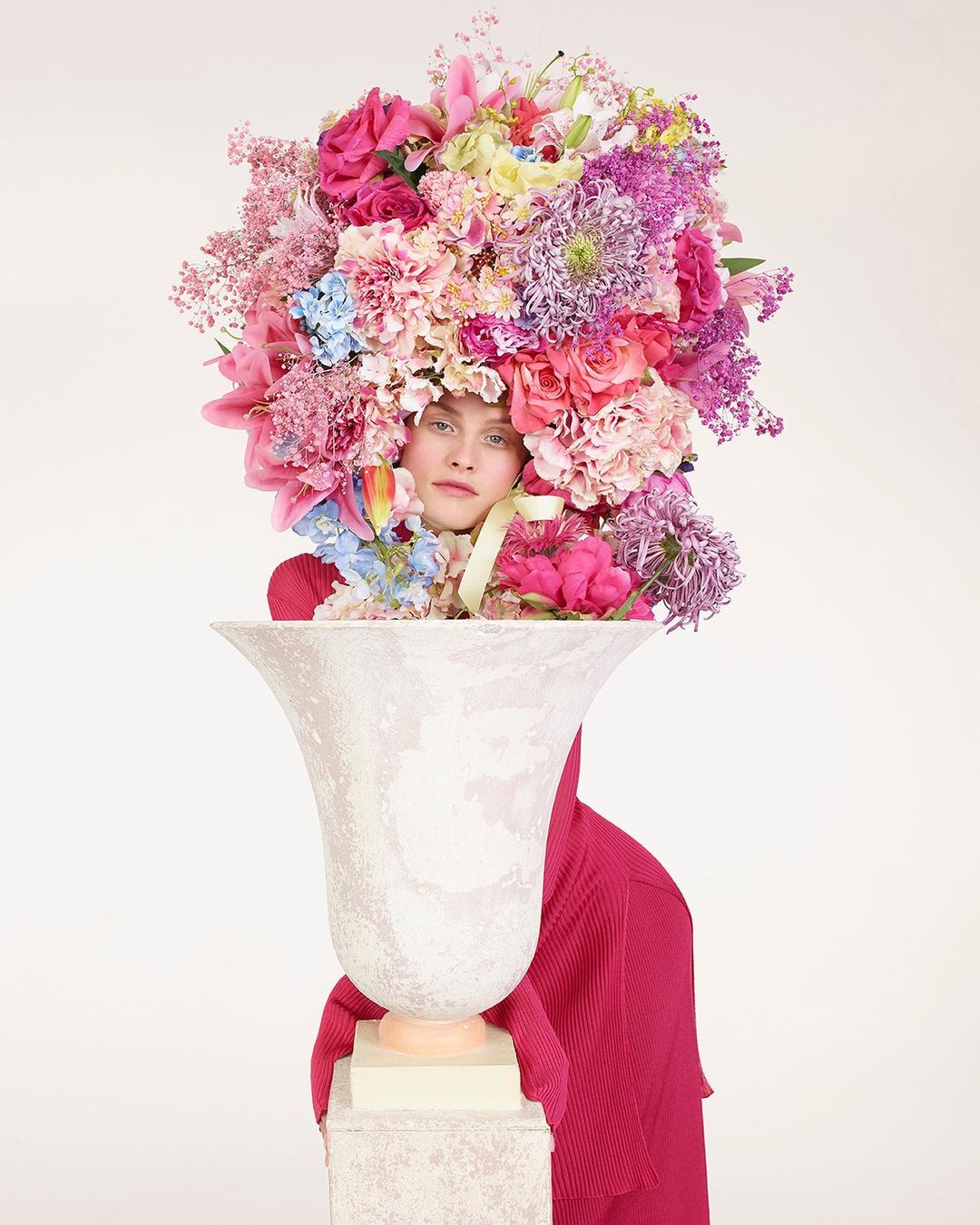 Woman stands behind a large decorative white vase, adorned with a colorful array of flowers. She is wearing a vibrant pink top.