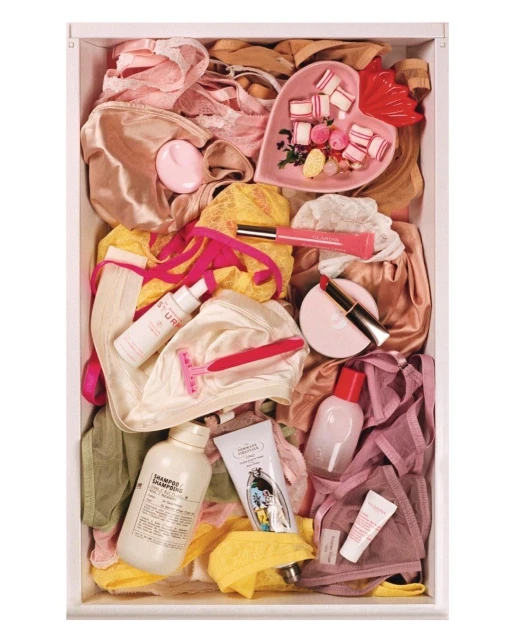 Overhead shot of a drawer filled with lingerie, skincare products, razors, makeup, and candy in a white drawer, creating a colorful still life.