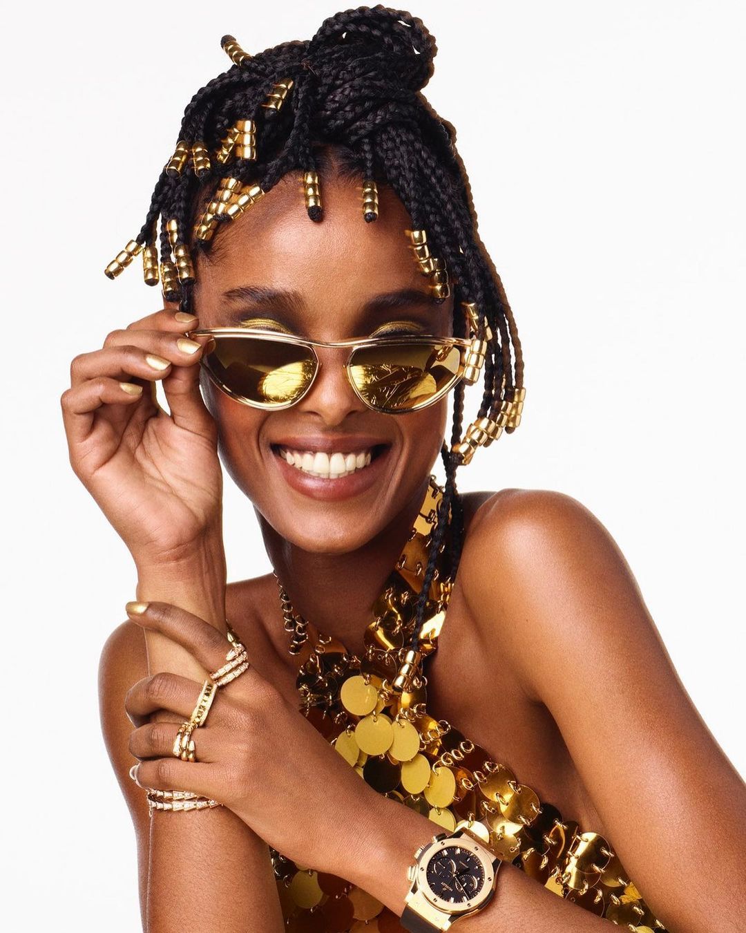 Smiling woman with dark hair in braids and gold beads, gold sunglasses, and a gold coin top against a white backdrop.