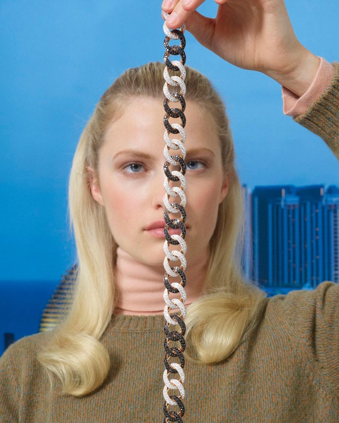 Woman presents chain-link bracelet with gem accents in a portrait against a blurred city skyline backdrop under a clear blue sky.