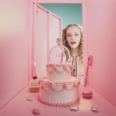 Chanel perfume bottle sits atop a two-tier cake next to a woman, macarons, cupcake, and candy cane in a monochromatic pink setting.