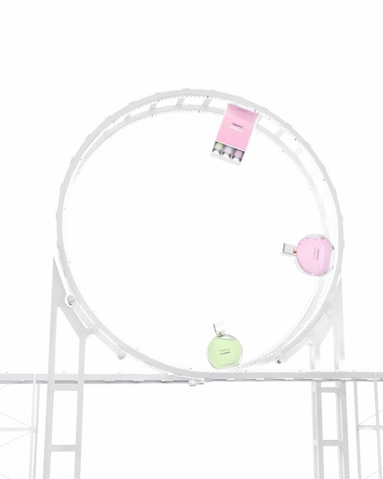 Three perfume bottles in shades of pink and green move on a white toy roller coaster track with a bright white backdrop. 