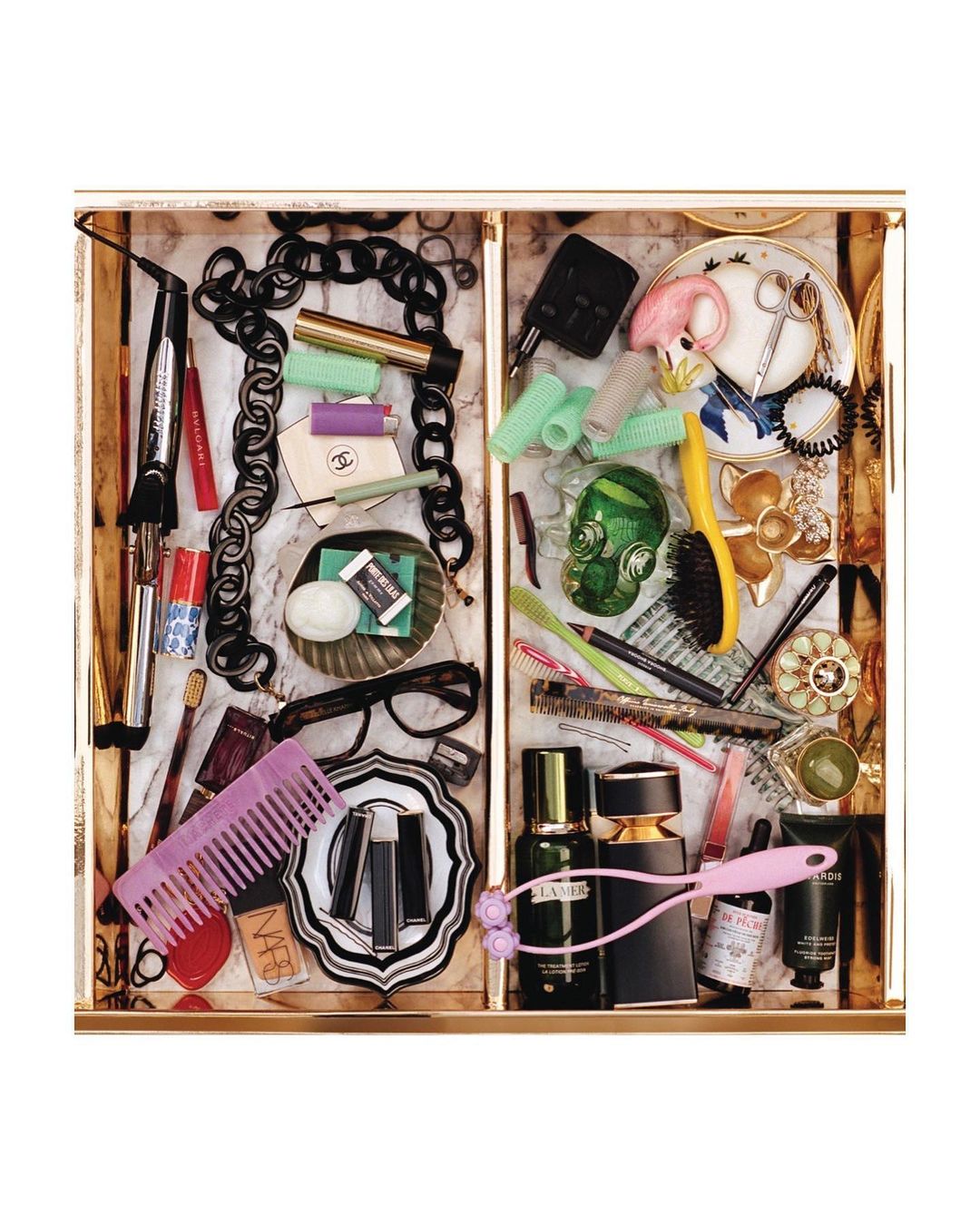 Overhead shot of an organized gold drawer containing beauty and makeup products: perfume bottles, cosmetics, hair accessories, brushes.