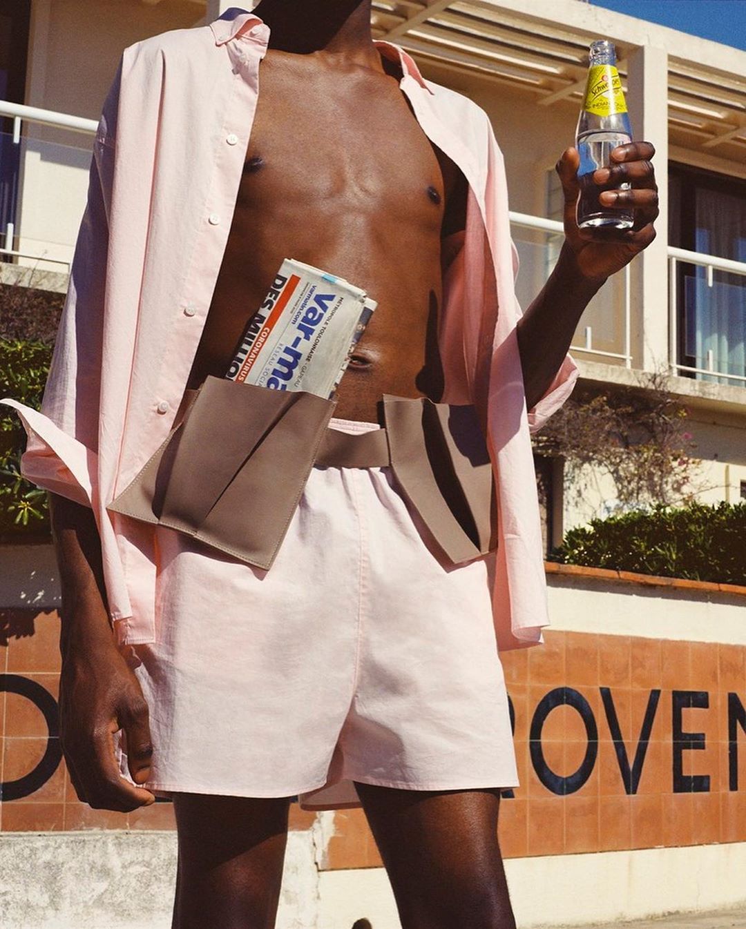 Fashion shot of a Black male model in pink shirt and shorts, holding a bottle and wearing a belt bag with newspaper, outside a building.