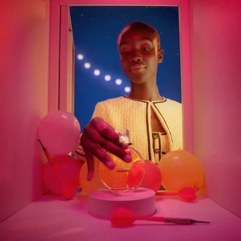 Elegant dark-skinned woman poses with Chanel perfume in an indoor studio setting with party balloons and decorative lighting.