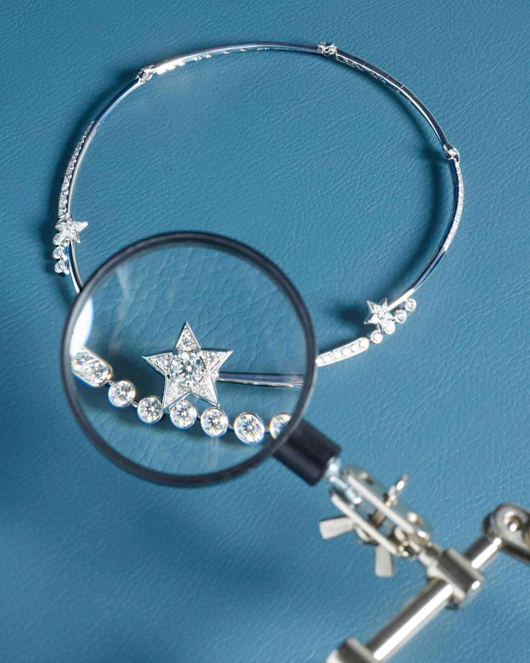 Sparkling diamond star choker necklace on blue surface being examined with a small magnifying glass and articulated tool arm.