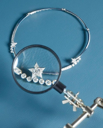 Sparkling diamond star choker necklace on blue surface being examined with a small magnifying glass and articulated tool arm.