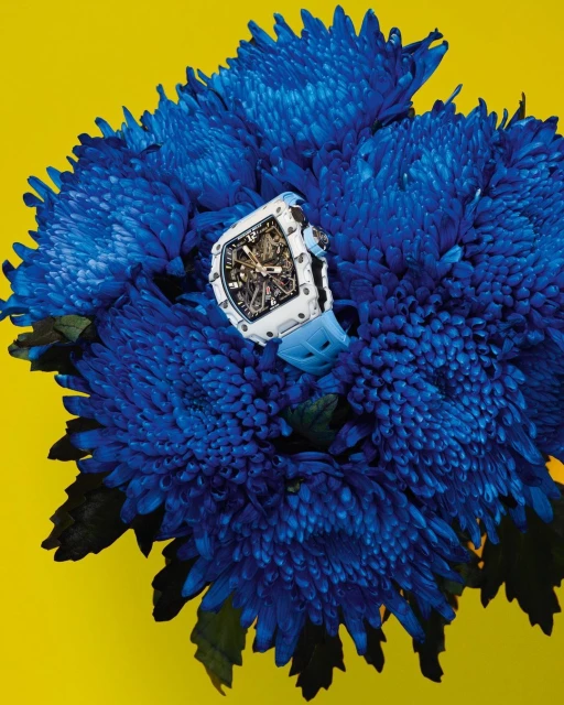 Luxury white wristwatch with a blue band displayed among vibrant blue chrysanthemum flowers against a bright yellow backdrop in a studio setting.